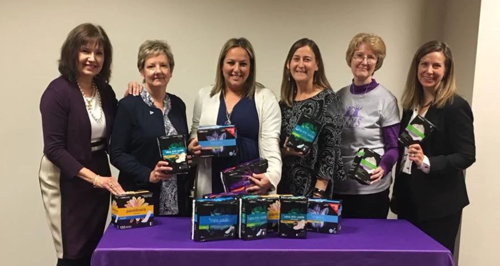 BRAWS Benefit Evening 2 Six women stand behind a table displaying menstrual hygiene products, smiling during a community donation and awareness event.
