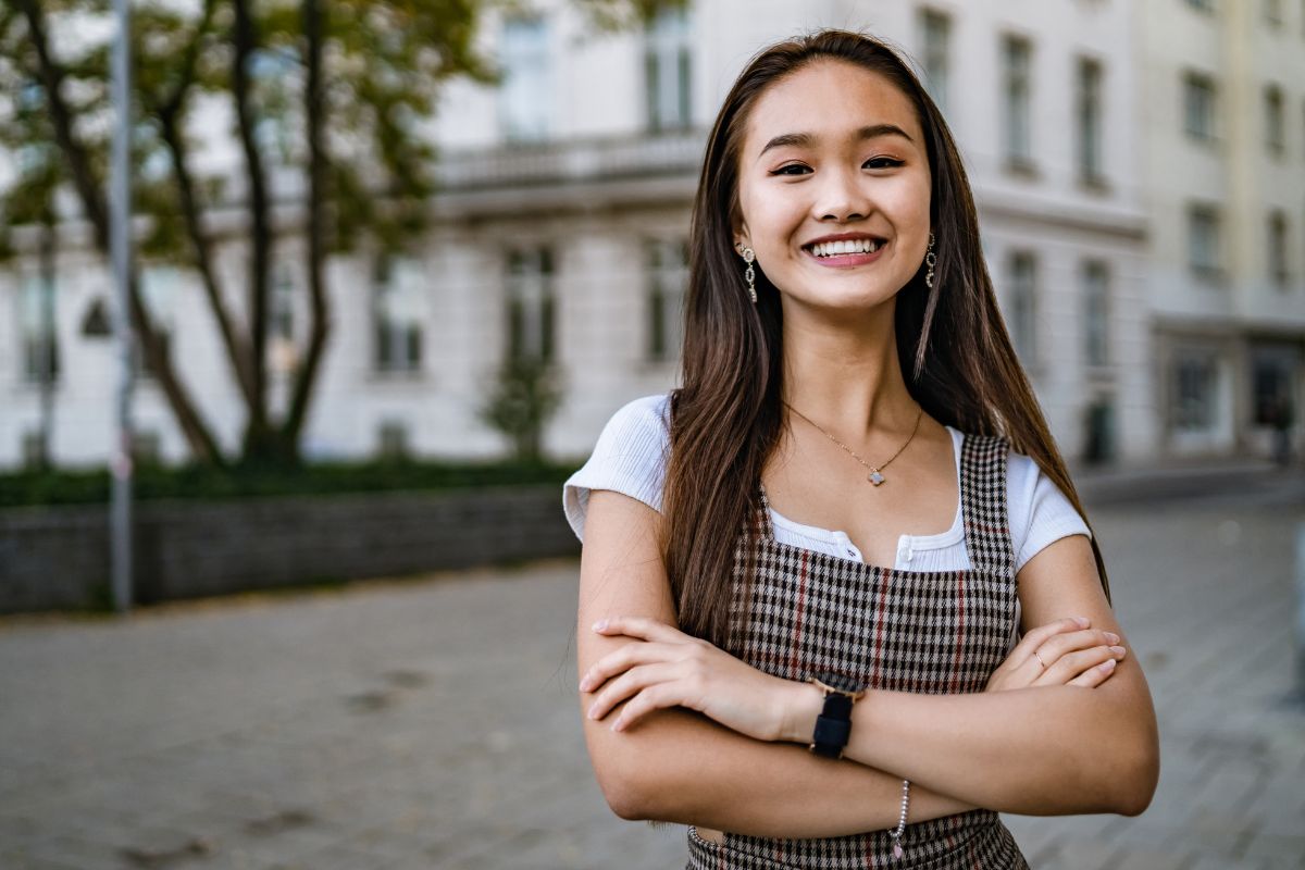 LCWAG Home Page Research & Impact Image depicting a young Asian woman with her arms crossed smiling in a city setting.